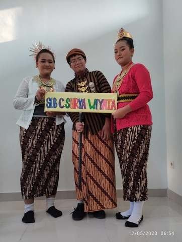 Guru dan siswi berkebaya dan berkain batik membawa papan SLB C Surya Wiyata